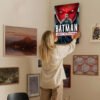 Person hanging Batman Under the Red Hood movie poster on eclectic gallery wall featuring various framed artworks in neutral-toned interior space.
