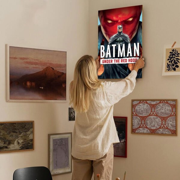 Person hanging Batman Under the Red Hood movie poster on eclectic gallery wall featuring various framed artworks in neutral-toned interior space.