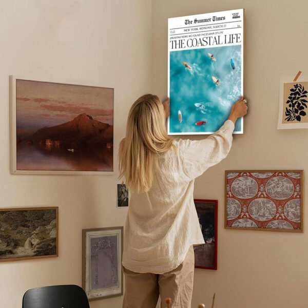 Woman styling gallery wall by hanging coastal life newspaper canvas art among eclectic collection of vintage and contemporary artworks in neutral home interior.