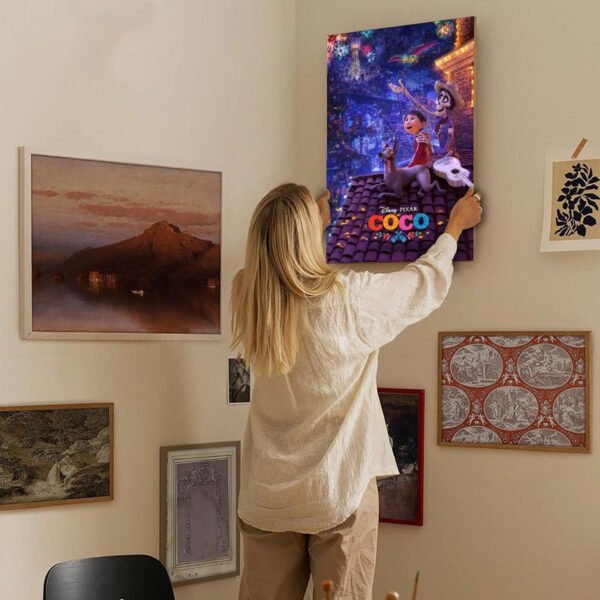 Person hanging Disney Pixar Coco movie poster on modern gallery wall with eclectic art collection including framed landscapes and botanical prints in minimalist home interior.