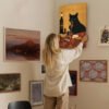 Woman arranging eclectic gallery wall featuring black cat wine art, landscape paintings, vintage maps, and botanical prints in modern home interior.