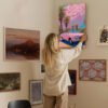 Woman arranging retro pink car canvas art on eclectic gallery wall showcasing home art curation and interior design styling techniques.