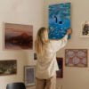 Woman arranging whimsical cat artwork on gallery wall in modern home interior with curated art collection.