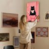 Woman hanging funny cat bathroom art print on eclectic gallery wall in modern home interior.