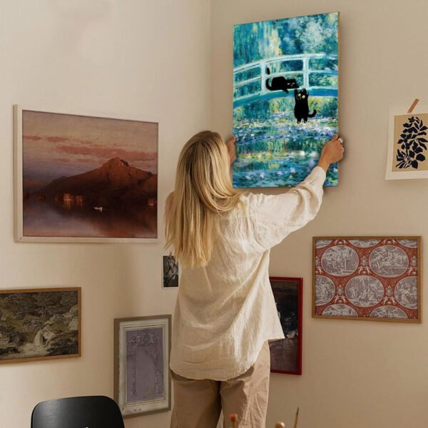 Woman hanging black cats impressionist artwork on curated gallery wall with mixed art collection in modern home interior.