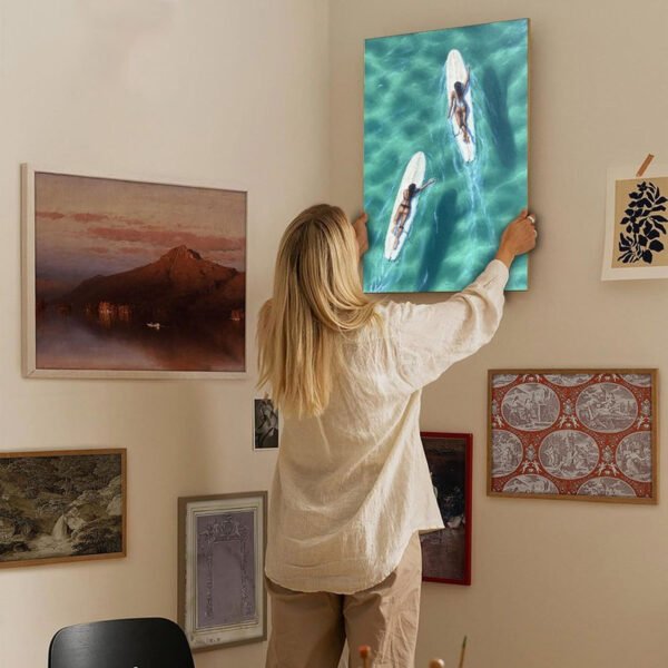 Woman hanging aerial surf canvas art on curated gallery wall featuring diverse framed artwork in modern home interior design setting.