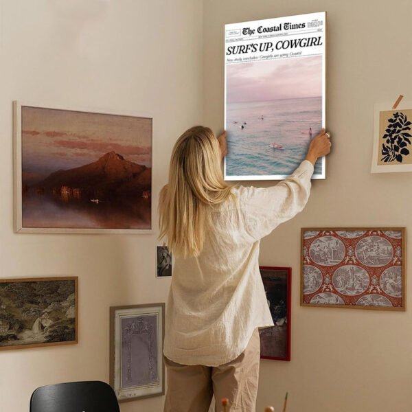 Woman styling gallery wall by hanging Coastal Times Surf's Up Cowgirl vintage newspaper canvas print among eclectic art collection