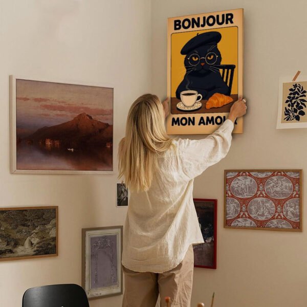 Woman in cream sweater hanging French "Bonjour Mon Amour" cat poster on curated gallery wall with various framed artworks in neutral-toned living space.
