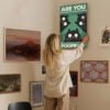Woman hanging funny cat poster "Are You Poopin'" on eclectic gallery wall with various framed artworks in modern home setting.