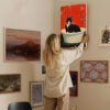 Woman styling a gallery wall by hanging vintage cat bathtub poster among curated art collection in modern home interior with eclectic decor.