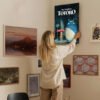 Woman hanging My Neighbor Totoro canvas print on eclectic gallery wall featuring various framed artwork in modern home interior.