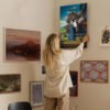 Person arranging Howl's Moving Castle poster on eclectic gallery wall featuring various framed artworks in modern home interior.