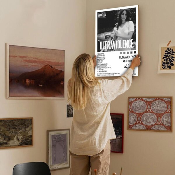 Person hanging Lana Del Rey Ultraviolence album poster on curated gallery wall with various framed artworks in modern home interior.
