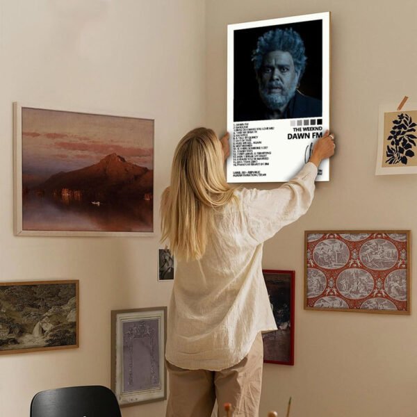 Person hanging The Weeknd Dawn FM poster on gallery wall with framed art in modern home interior