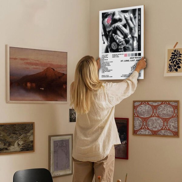 Person hanging A$AP Rocky album poster on gallery wall featuring eclectic art collection in modern home interior with warm neutral decor and natural lighting.