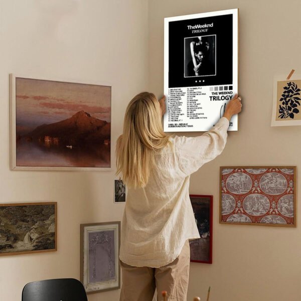 Young woman hanging The Weeknd Trilogy music poster on gallery wall in cozy bedroom with eclectic art collection and warm interior design.