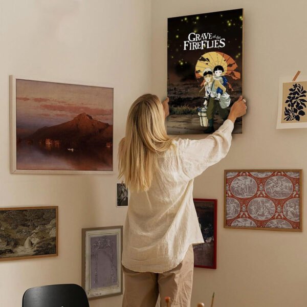 Person hanging Grave of the Fireflies anime poster on curated gallery wall with multiple framed artworks