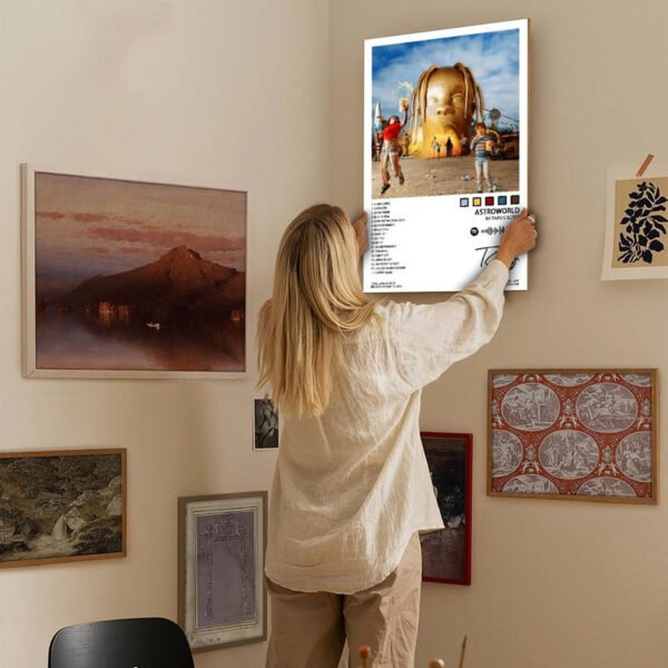 Person in cream sweater hanging Travis Scott ASTROWORLD poster on curated gallery wall featuring mix of landscape art, vintage maps, and decorative prints in warm neutral-toned interior space, demonstrating music poster home decor integration.
