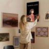 Person hanging Spirited Away canvas print on curated gallery wall with mixed artwork including landscapes and vintage maps.