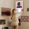 Woman hanging Tyler The Creator IGOR album poster with pink background and track listing on gallery wall among framed artworks in home interior