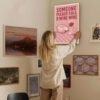 Woman hanging humorous wine-themed wall art "Someone Please Call 9 Wine Wine" on curated gallery wall with vintage maps and landscape paintings.