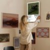 music-poster-gallery-wall-home-decor-styling.jpg Woman hanging Pierce The Veil album poster on eclectic gallery wall featuring various framed artworks in cozy home interior